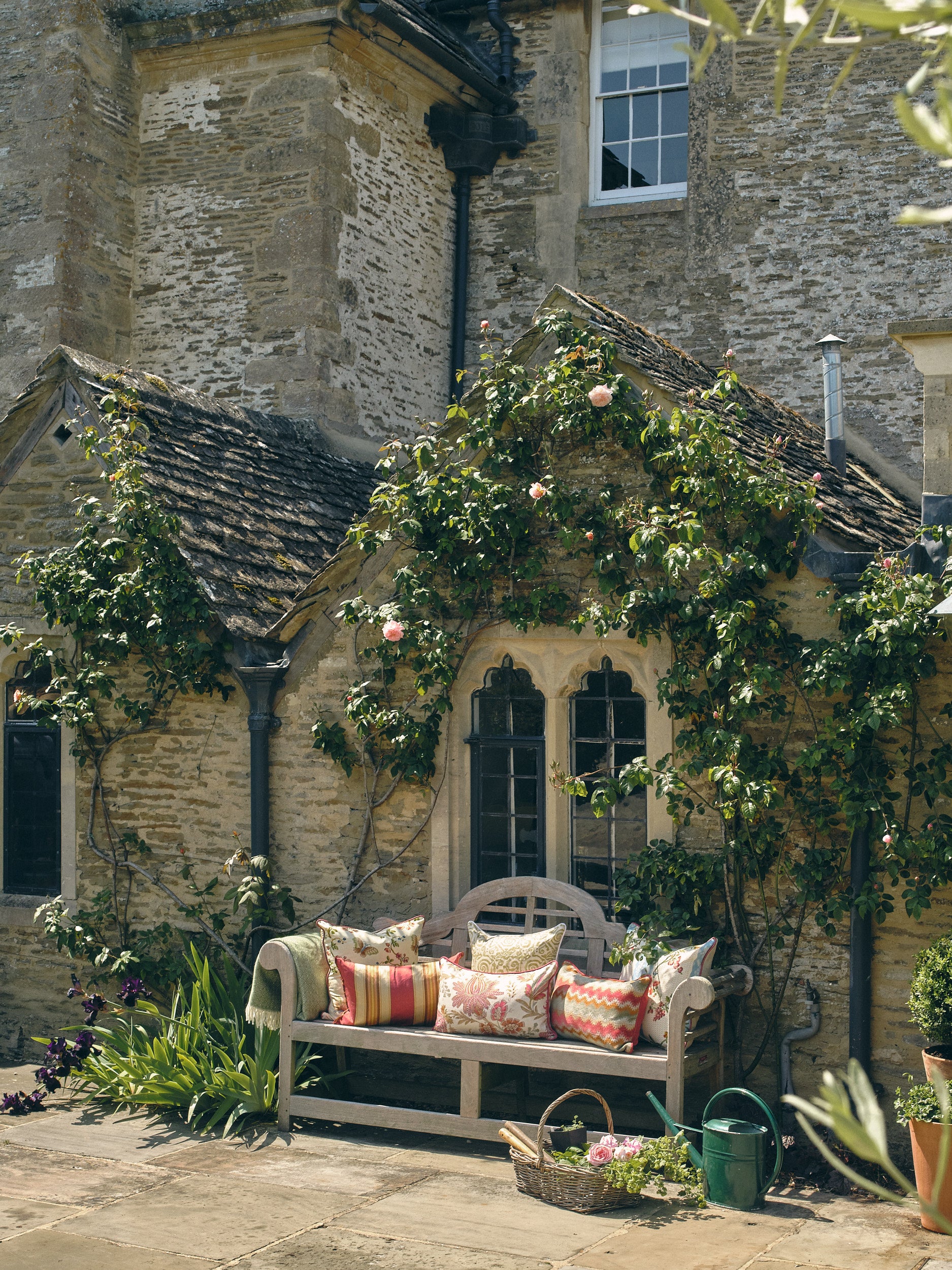Wooden bench with floral cushions in front of a stone building with climbing plants.