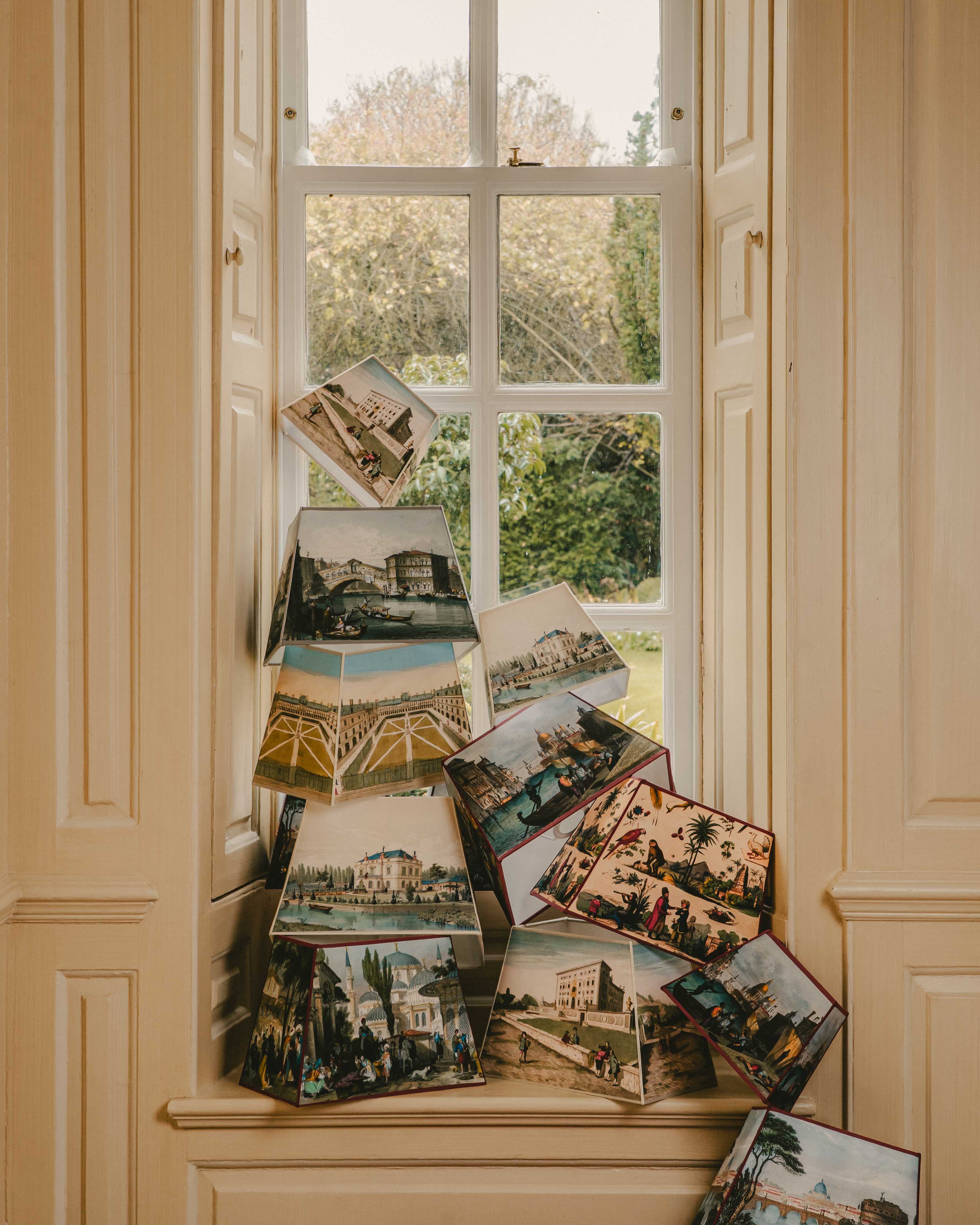 Stack of vintage looking lampshades on a window sill with a view of greenery outside.