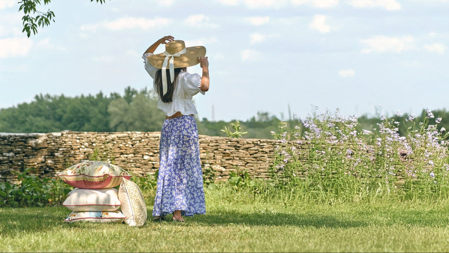 Woman in a long dress standing in a field with a stone wall and trees in the background